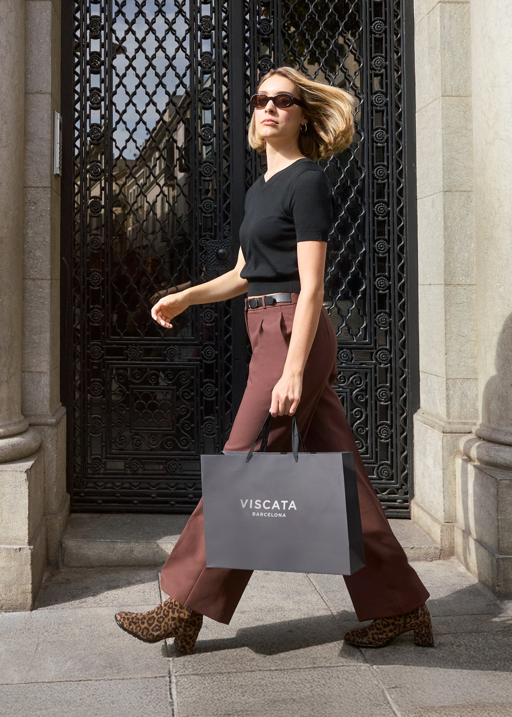 Woman walking confidently outdoors wearing black top and brown trousers, carrying a Viscata Barcelona shopping bag and styled in leopard print ankle boots — Viscata Essenza street style look.