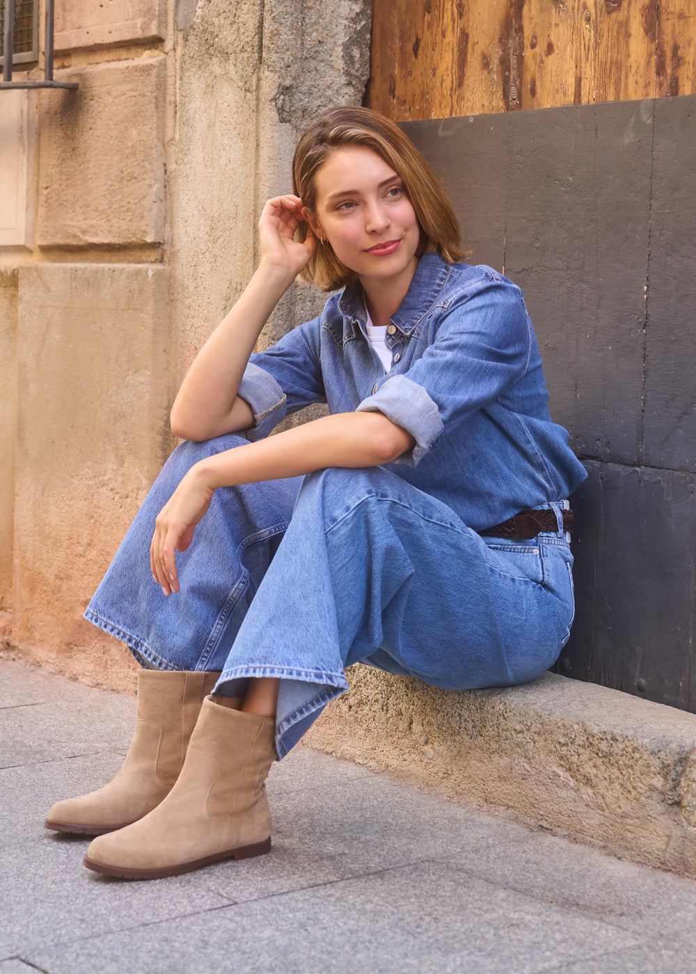 Woman sitting on a stone ledge wearing a denim outfit and camel suede ankle boots, smiling casually — Viscata Cremona boots street-style look.