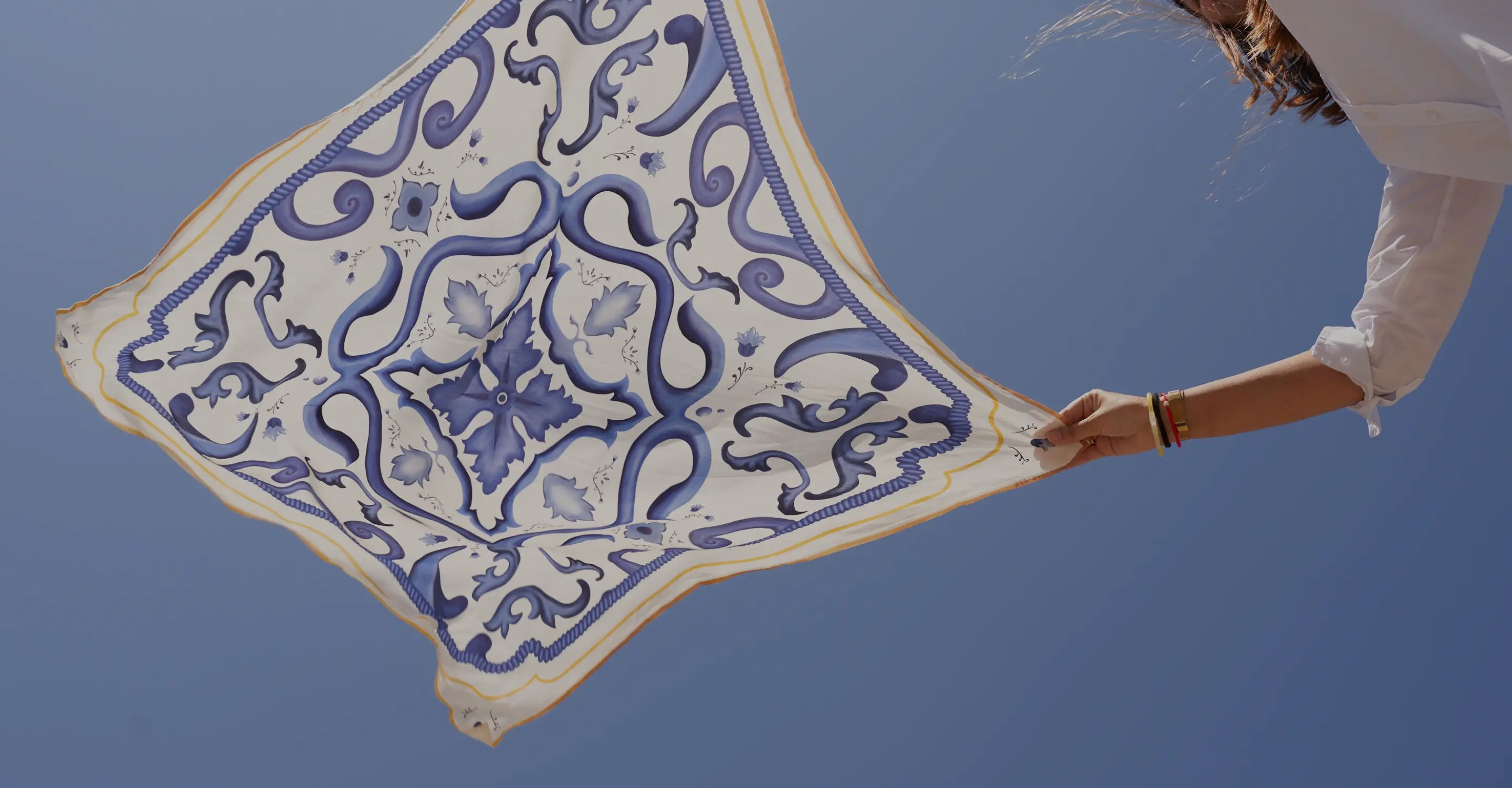 Woman holding a blue and white Mediterranean tile-print silk scarf against a clear blue sky, showcasing elegant summer accessories — perfect for coastal vacations, lightweight layering, and European-inspired fashion