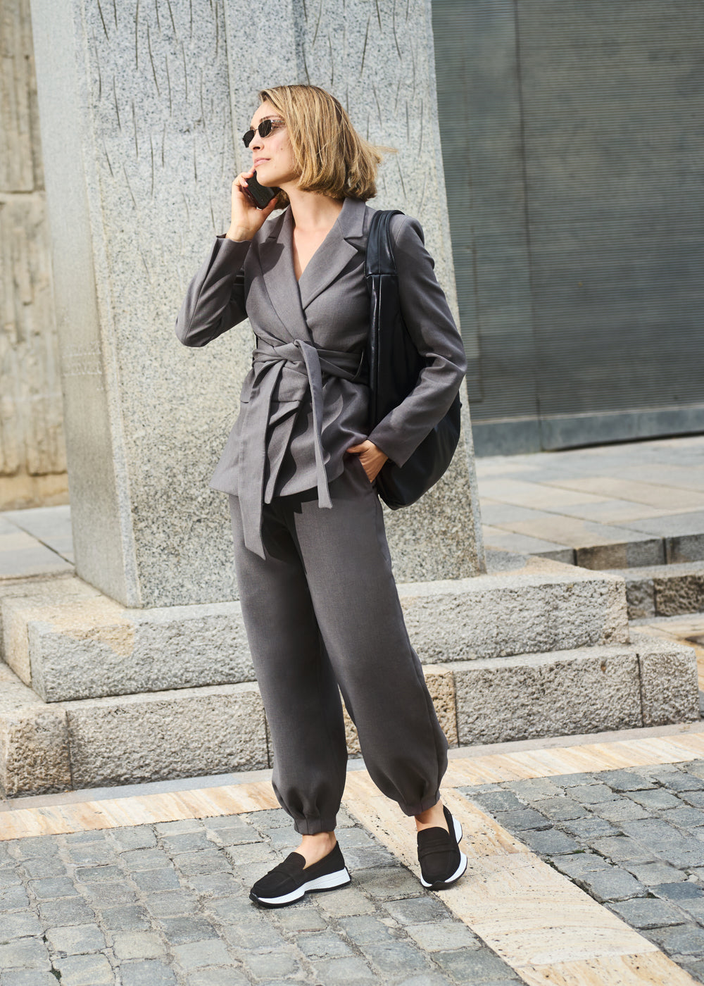 Woman talking on the phone while walking on a city street, wearing a grey belted suit and Modena black slip-on loafers with white soles — Viscata casual-chic look.