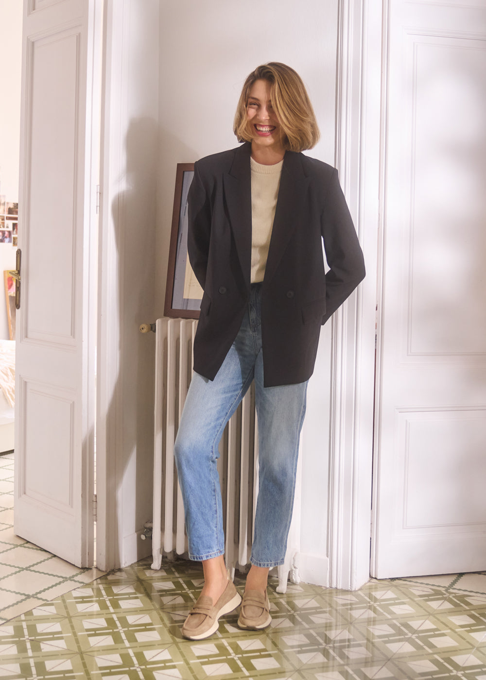 Woman smiling indoors wearing light sweater, black blazer, and light blue jeans paired with camel suede loafers — Viscata Modena casual chic lifestyle look.