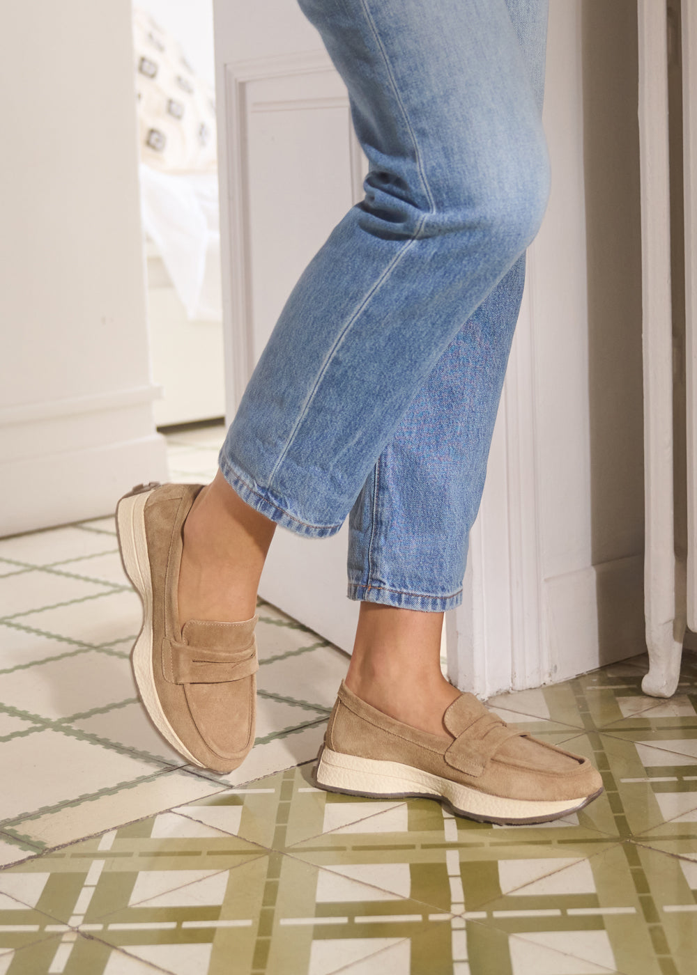 Close-up of camel suede slip-on loafers with white soles, styled with cropped blue jeans indoors — Viscata Modena shoes detail shot.