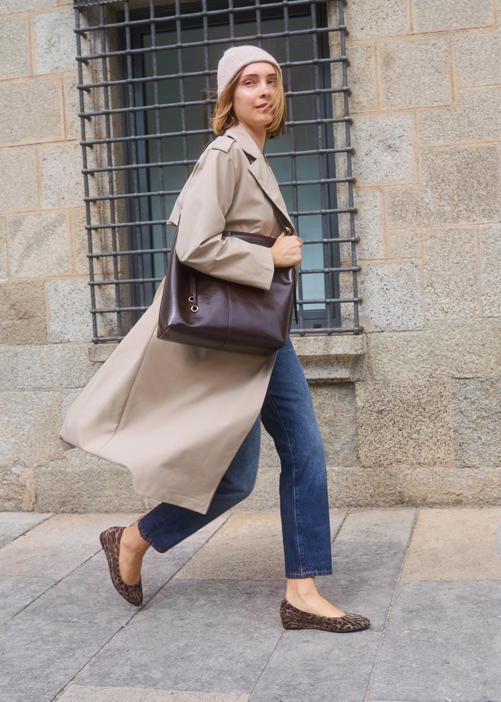 Woman walking down the street wearing a beige trench coat, jeans, and leopard-print flats, carrying a dark brown leather handbag and beige knit beanie — Viscata Roma flats urban fall look.