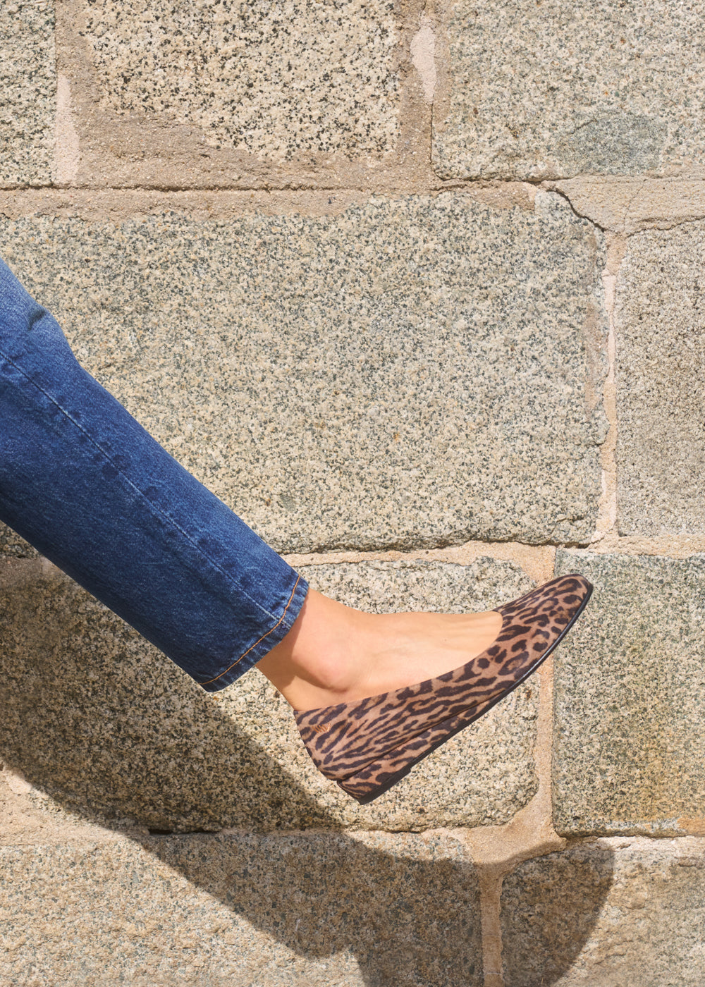 Close-up of a woman’s foot wearing leopard-print flat suede shoes paired with blue jeans against a stone wall — Viscata Roma flats.