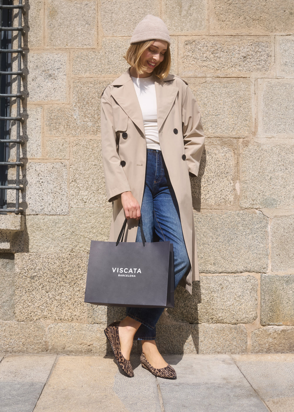 Woman wearing a beige trench coat, white top, jeans, and Roma leopard-print suede flats, holding a Viscata Barcelona shopping bag while standing against a stone wall.