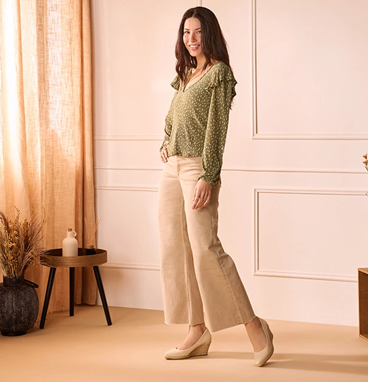 Woman smiling while modeling Viscata beige medium wedge espadrilles, paired with wide-leg beige pants and a green patterned blouse, standing in a softly lit room with neutral decor.