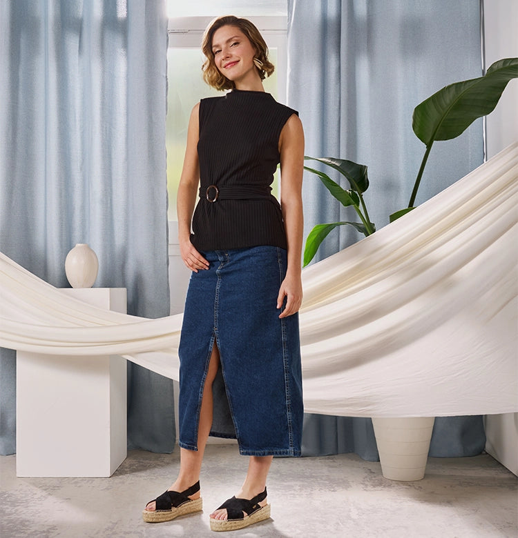 Woman standing indoors wearing a sleeveless black top with a belt, a high-slit denim midi skirt, and black espadrille platform sandals with knotted straps, posing in front of light blue curtains and a white draped backdrop.
