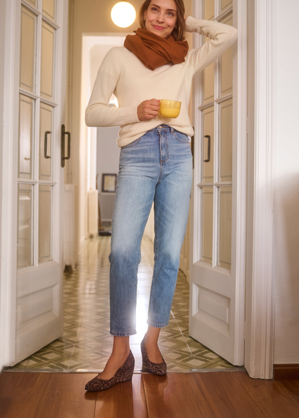 Woman standing in a cozy home setting holding a yellow mug, wearing light blue jeans, cream sweater, and brown scarf, styled with leopard-print wedges— Viscata Verona wedges lifestyle look.