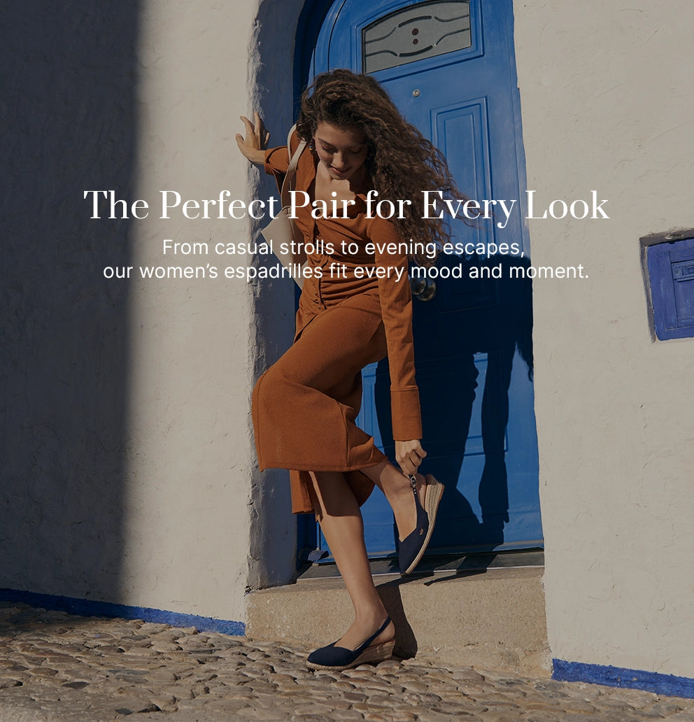 Woman wearing burnt orange outfit and navy slingback espadrille wedges, leaning playfully against a blue door on a cobblestone street