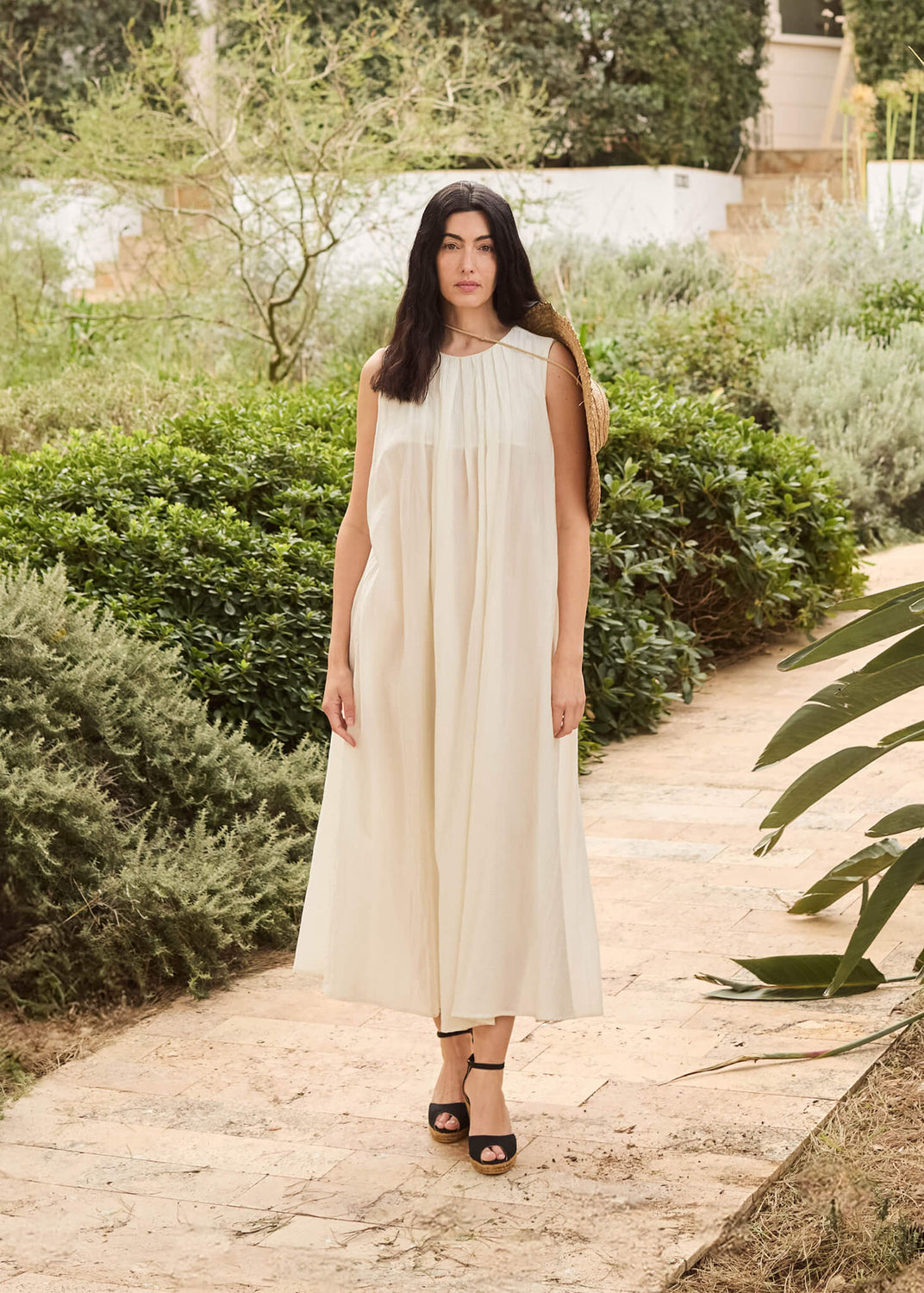 Woman standing along a garden path surrounded by lush greenery, wearing a flowing cream dress and Aiguafreda black ankle-strap wedge espadrilles. A calm, sunlit scene that reflects effortless Mediterranean grace.