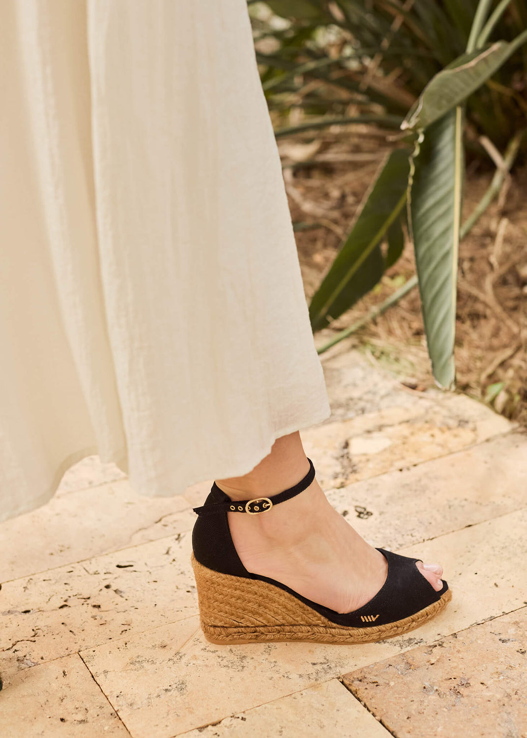Close-up of a woman wearing Aiguafreda black canvas ankle-strap wedge espadrilles, paired with a flowing cream dress, standing on a sunlit stone terrace surrounded by greenery.