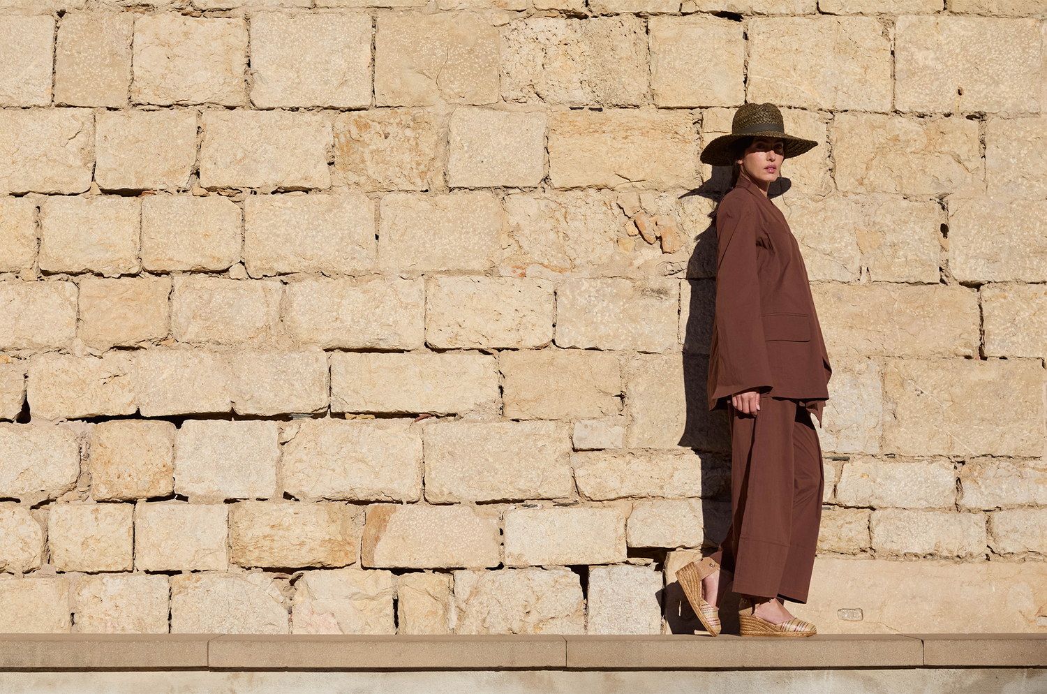 Person in brown outfit and hat standing against a stone wall