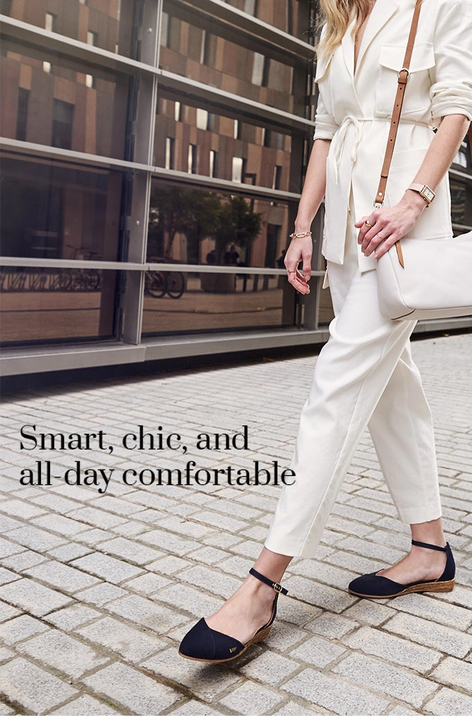 Viscata woman in white tailored suit walking outdoors, styled with navy ankle-strap flat espadrilles. Smart, chic, and all-day comfortable footwear for a modern lifestyle