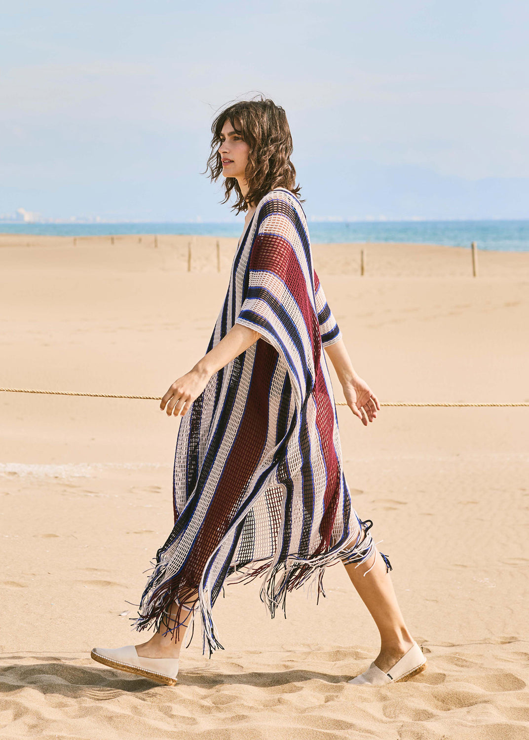 Woman walking on the beach wearing beige canvas slip-on espadrille flats, styled with a striped knit cover-up in a Mediterranean coastal setting.