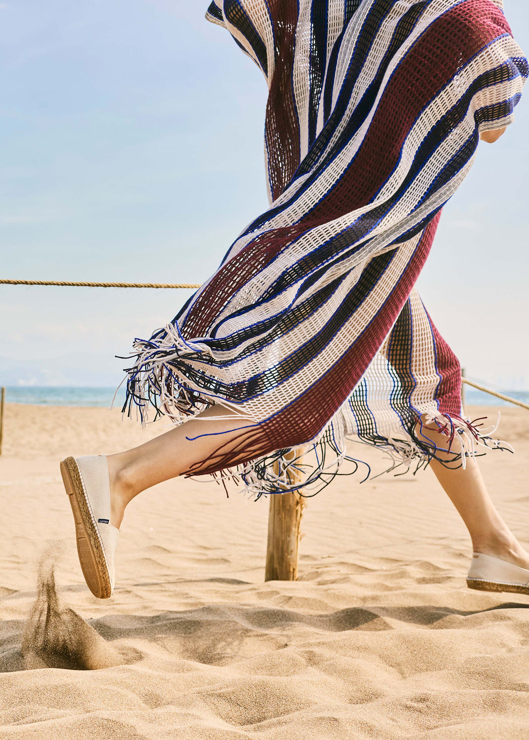Woman walking barefoot-inspired on the beach wearing beige canvas slip-on espadrille flats, styled with a flowing striped knit cover-up in motion.
