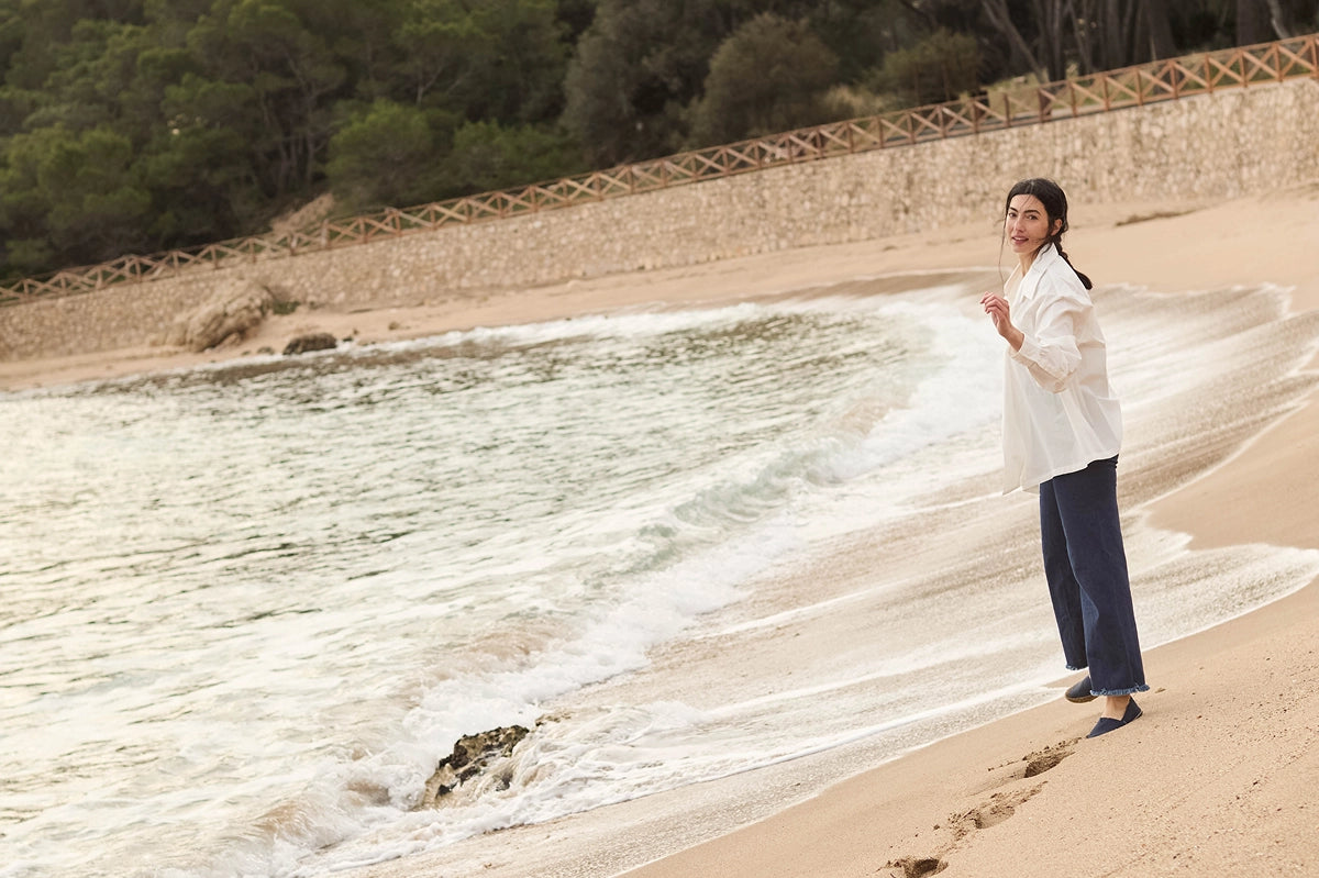 A woman stands on a sandy beach at the shoreline, wearing a loose white long-sleeve top and dark navy wide-leg pants. Waves are visible in the water, with a stone wall and green trees in the background.