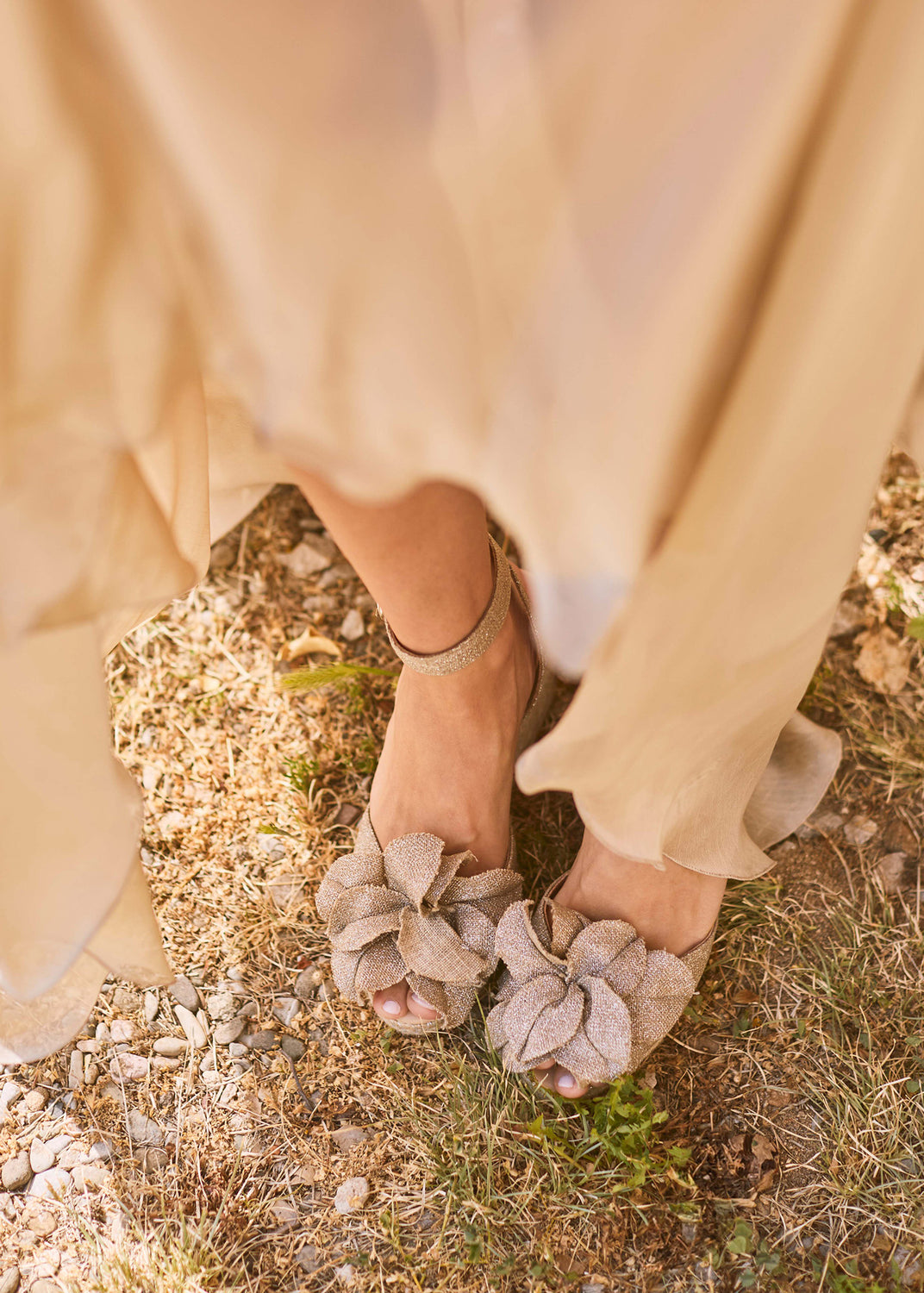 Champagne metallic espadrille wedge sandals with oversized bow detail styled beneath a flowing neutral dress outdoors.