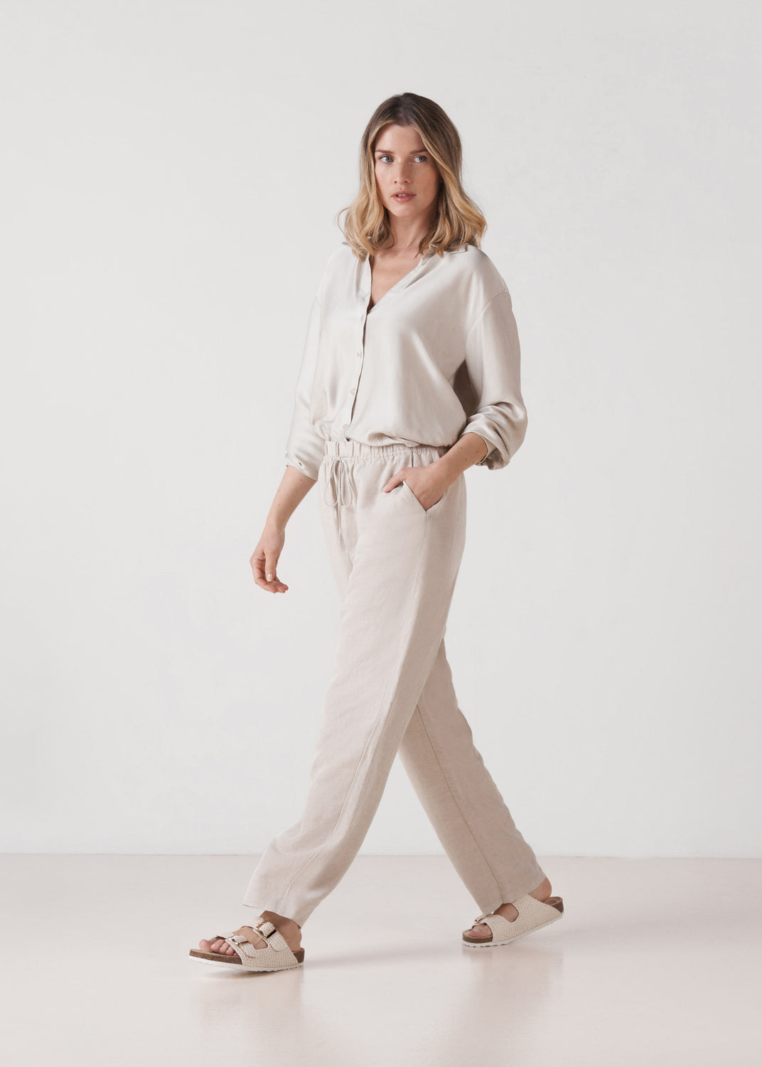 Model walking wearing natural raffia double-buckle slide sandals.