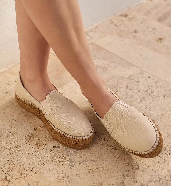 A person wearing off-white slip-on espadrille-style shoes with woven cork wedge soles, standing on a light beige stone surface.