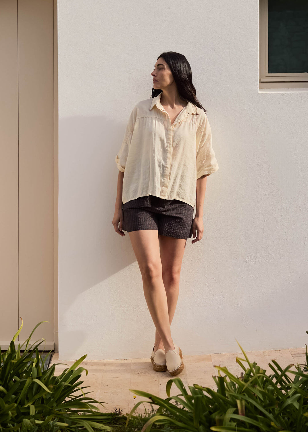 Woman standing against a white wall surrounded by greenery, wearing a loose beige shirt, dark shorts, and Castell beige canvas slip-on flats, embodying relaxed Mediterranean ease.