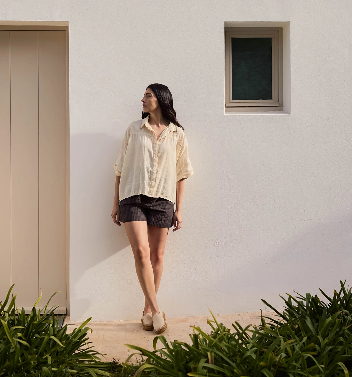 A woman stands outdoors against a white stucco wall, wearing a loose cream short-sleeve button-up shirt and dark shorts. The scene includes a doorway on the left, a small window near the top right, and green plants in the foreground.
