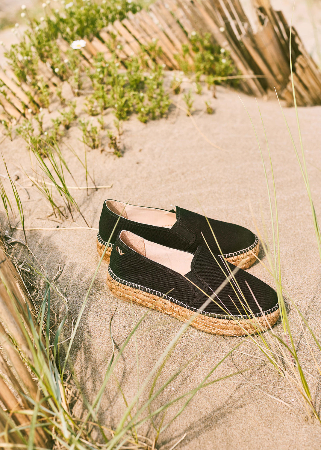 Black slip-on espadrille flats with braided jute soles placed on Mediterranean sand dunes in warm natural light.