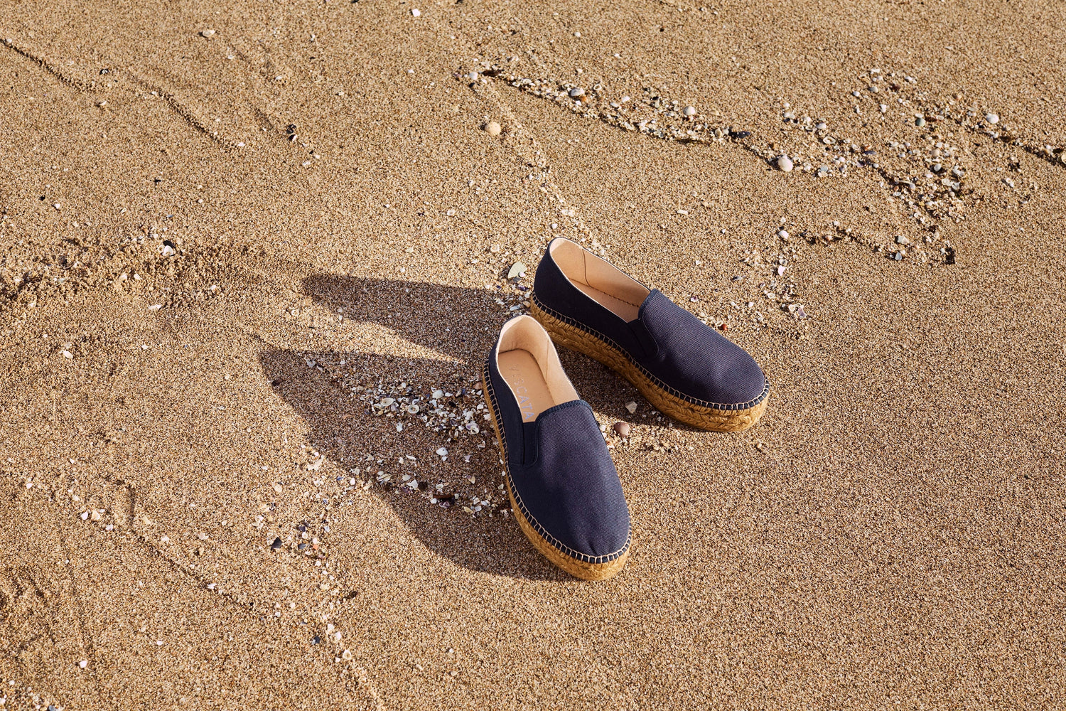A pair of navy blue slip-on espadrille-style shoes with tan soles, placed on sandy beach ground with shell fragments.