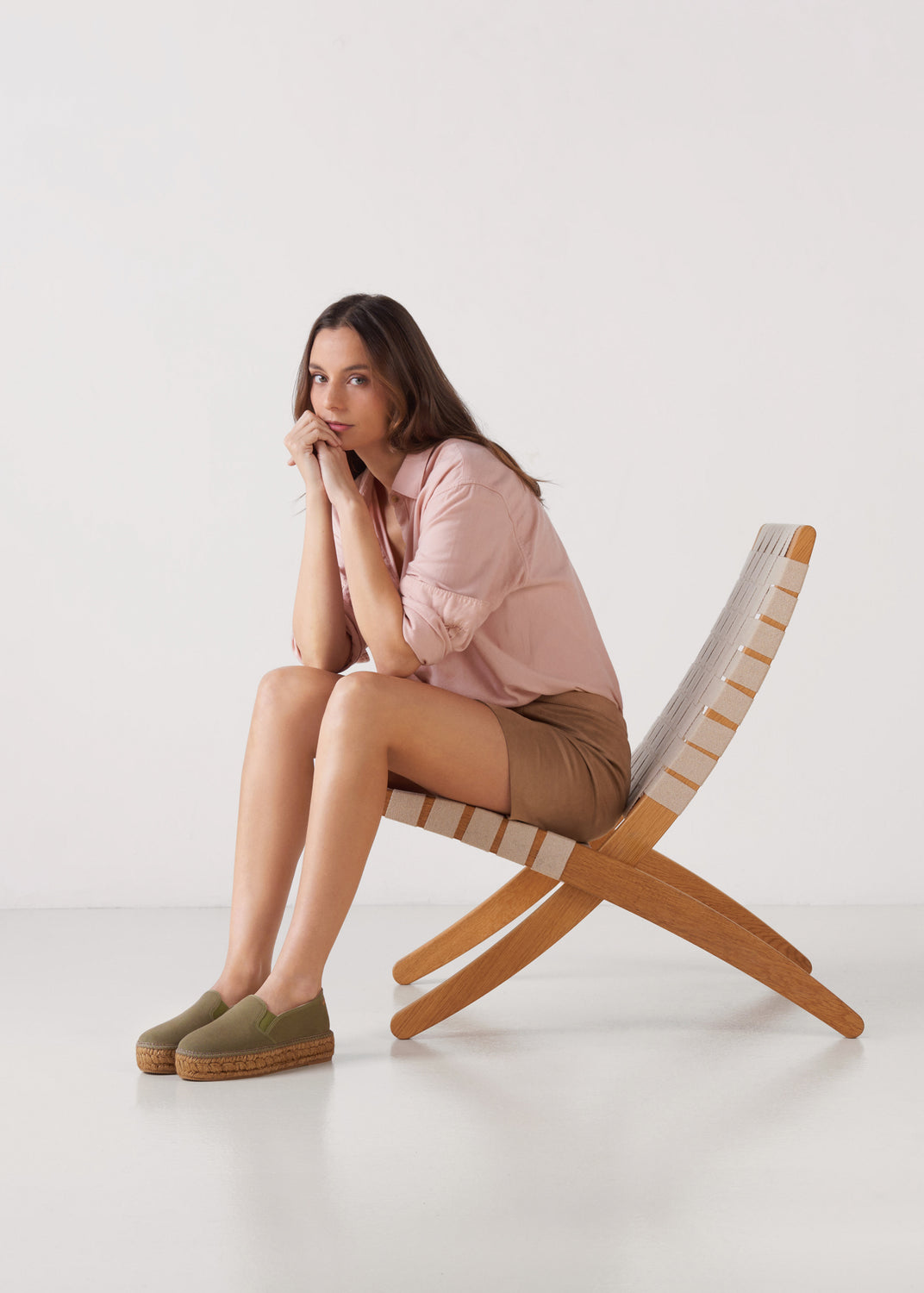 Woman seated in a woven lounge chair wearing olive green espadrille flats styled with a blush shirt and tan shorts in a clean studio setting