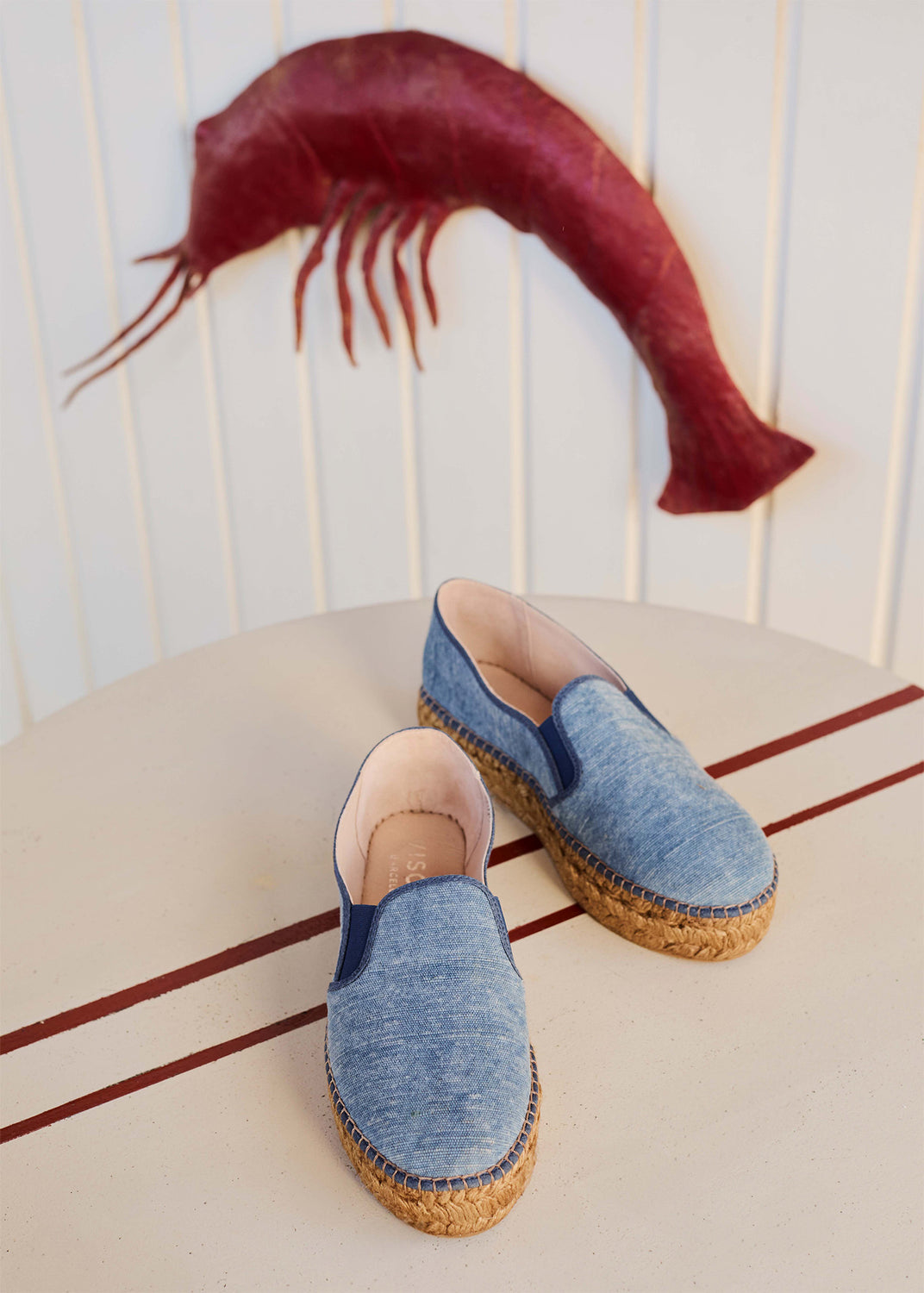 Blue denim espadrille slip-ons with jute sole placed on a table, with a red shrimp artwork in the background.