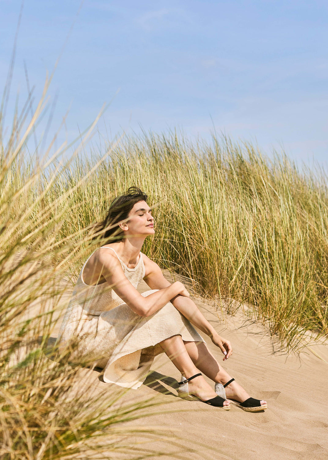 Woman relaxing on Mediterranean sand dunes wearing black espadrille wedge sandals with ankle straps and braided jute soles.