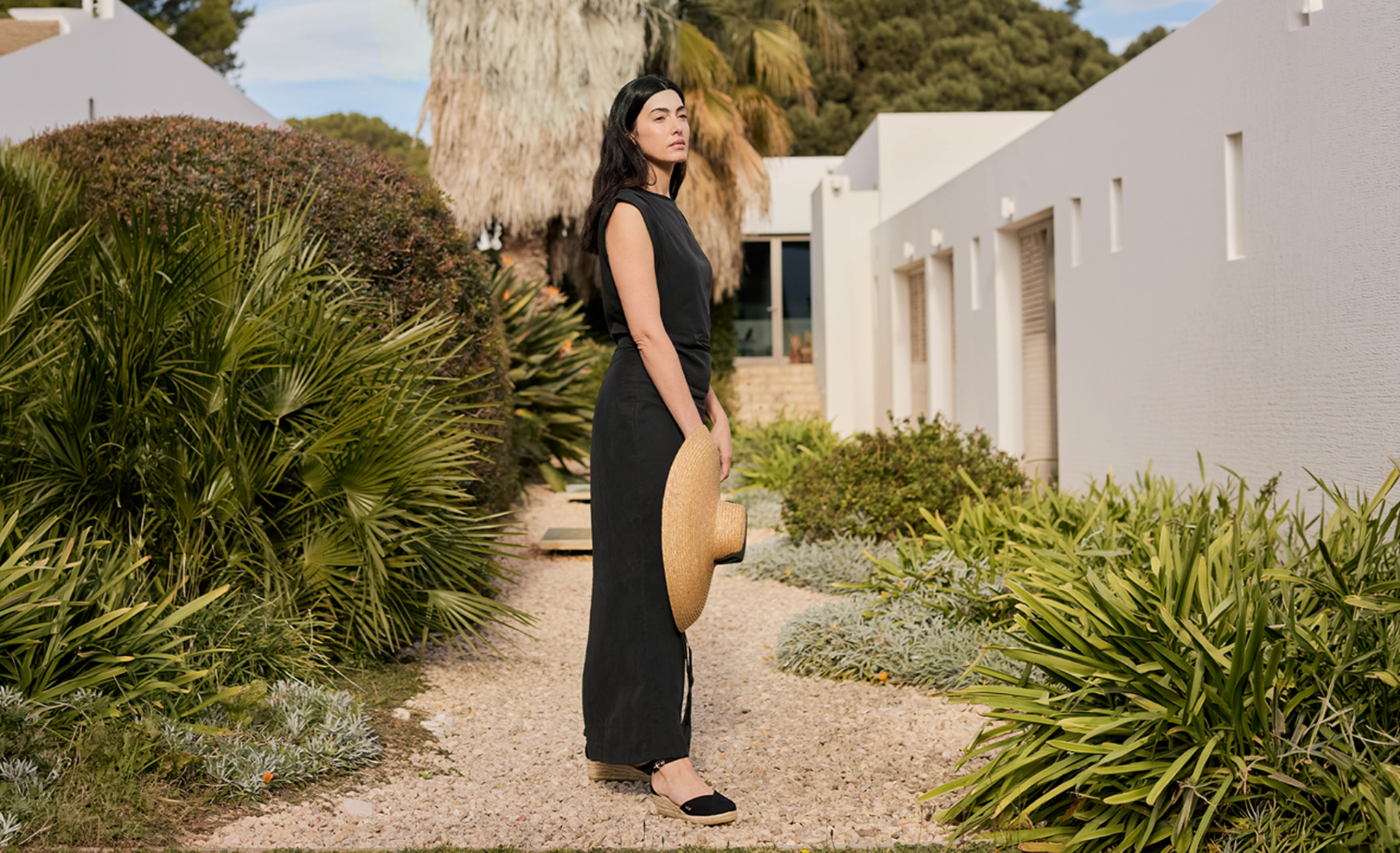 Woman in a black dress wearing black espadrille wedges, standing in a Mediterranean garden with warm natural light.