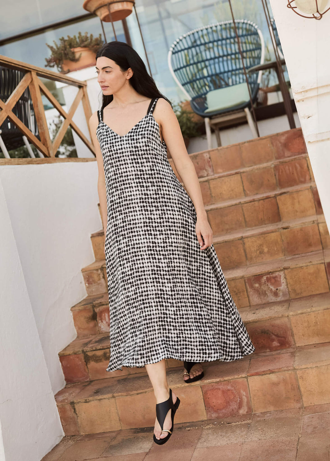 Woman walking down terracotta steps in a flowing black-and-white check dress, wearing Delmar black leather strap flats. A relaxed yet refined moment of Mediterranean style and ease.