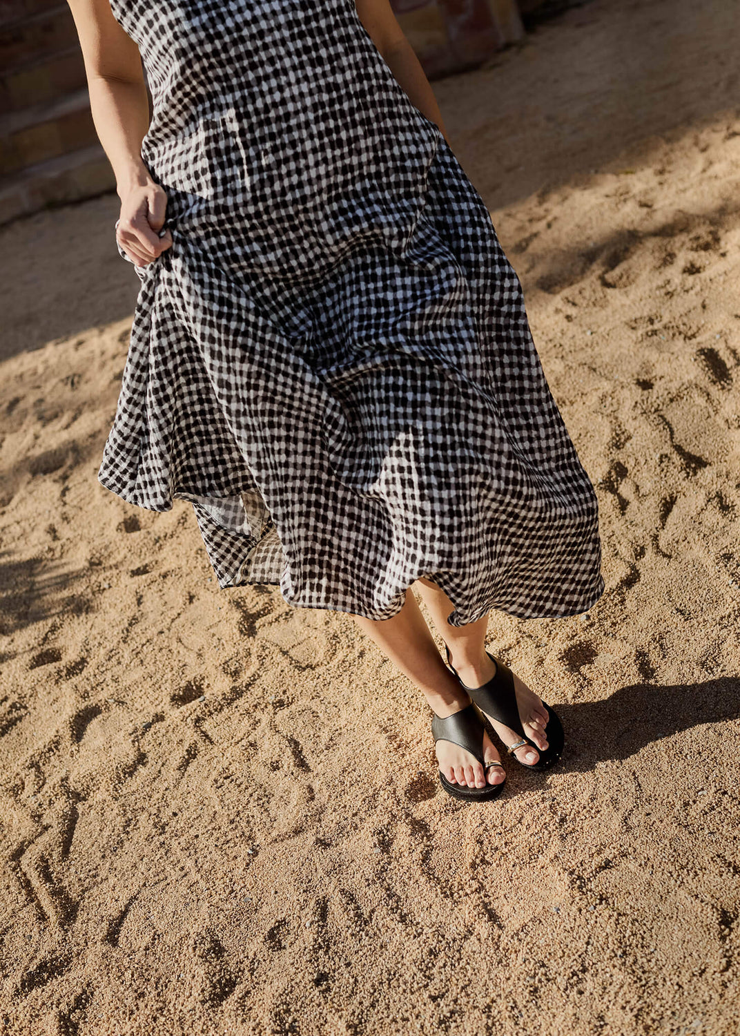 Close-up of a woman walking across sunlit sand in a flowing gingham dress and Delmar black leather strap espadrilles. A balance of effortless movement and timeless Mediterranean elegance.
