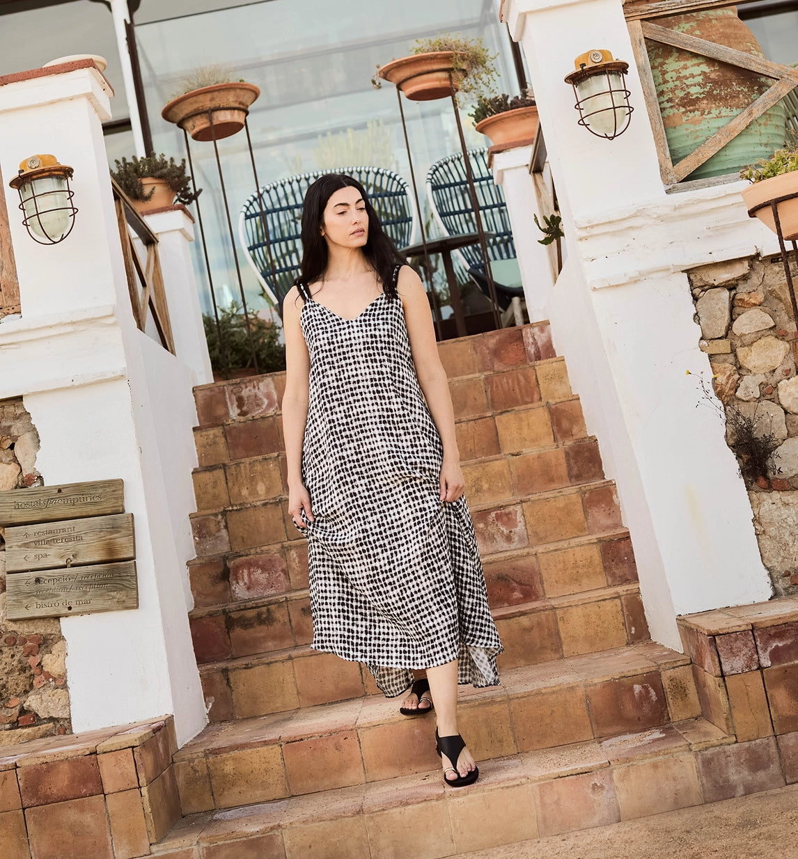 A woman wearing a black and white houndstooth midi dress stands on outdoor stone steps between white pillars with wall lanterns.