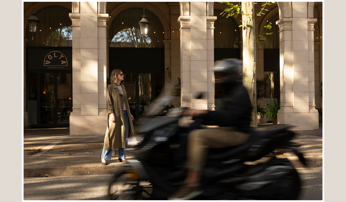 A woman in a long beige trench coat and blue jeans stands in a sunlit urban arcade with stone columns, while a blurred motorcyclist passes in the foreground.