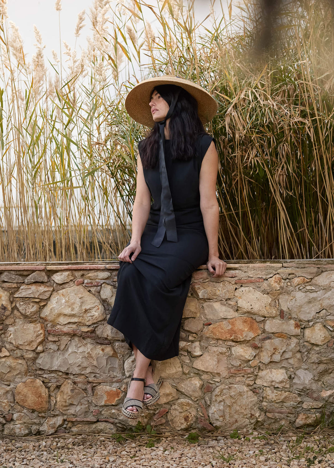 Woman sitting on a rustic stone wall surrounded by tall golden reeds, wearing a sleeveless black dress, wide straw hat, and Empuria black ankle-strap wedge espadrilles. A relaxed coastal mood with understated sophistication.