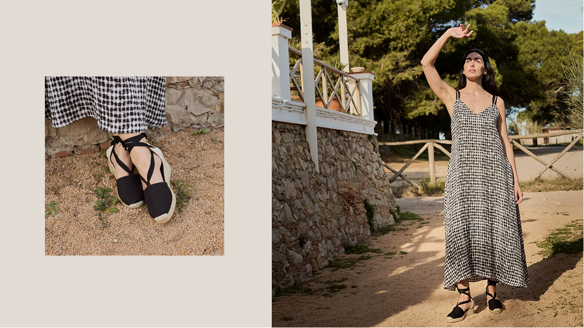 Woman in a checkered dress and black sandals standing outdoors with a stone wall and trees in the background.