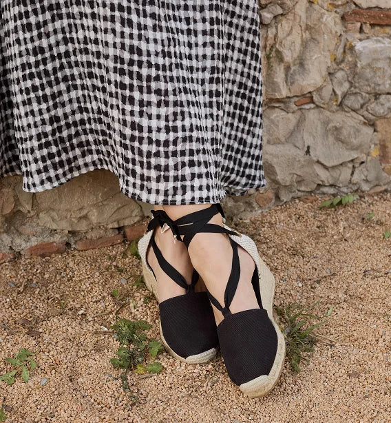 A person wearing black strappy wedge espadrille sandals with woven jute soles, standing outdoors on sandy ground in front of a stone wall.