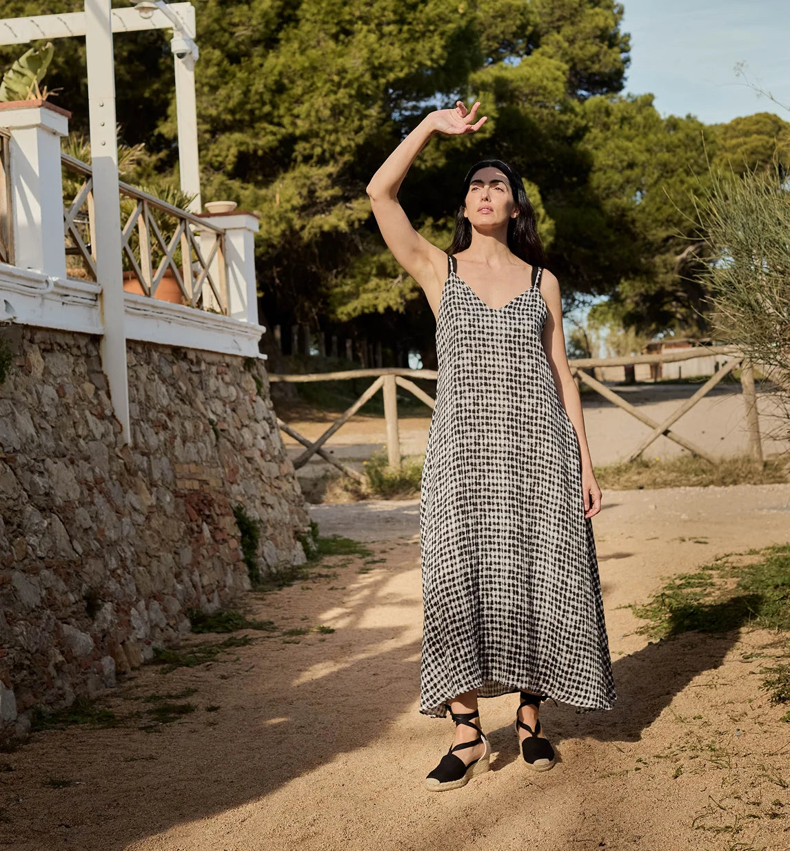 A woman wearing a black and white houndstooth midi dress and black espadrille sandals stands on a dirt path outdoors, with trees, a stone wall, and a white wooden structure in the background.
