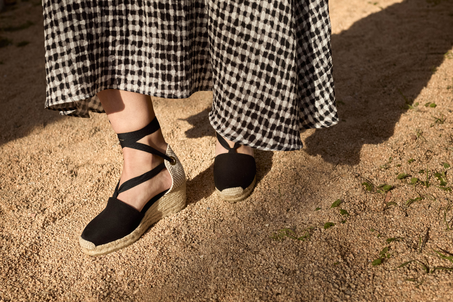 Black espadrille wedges with ankle ties worn outdoors with a gingham dress, styled in a Mediterranean setting.