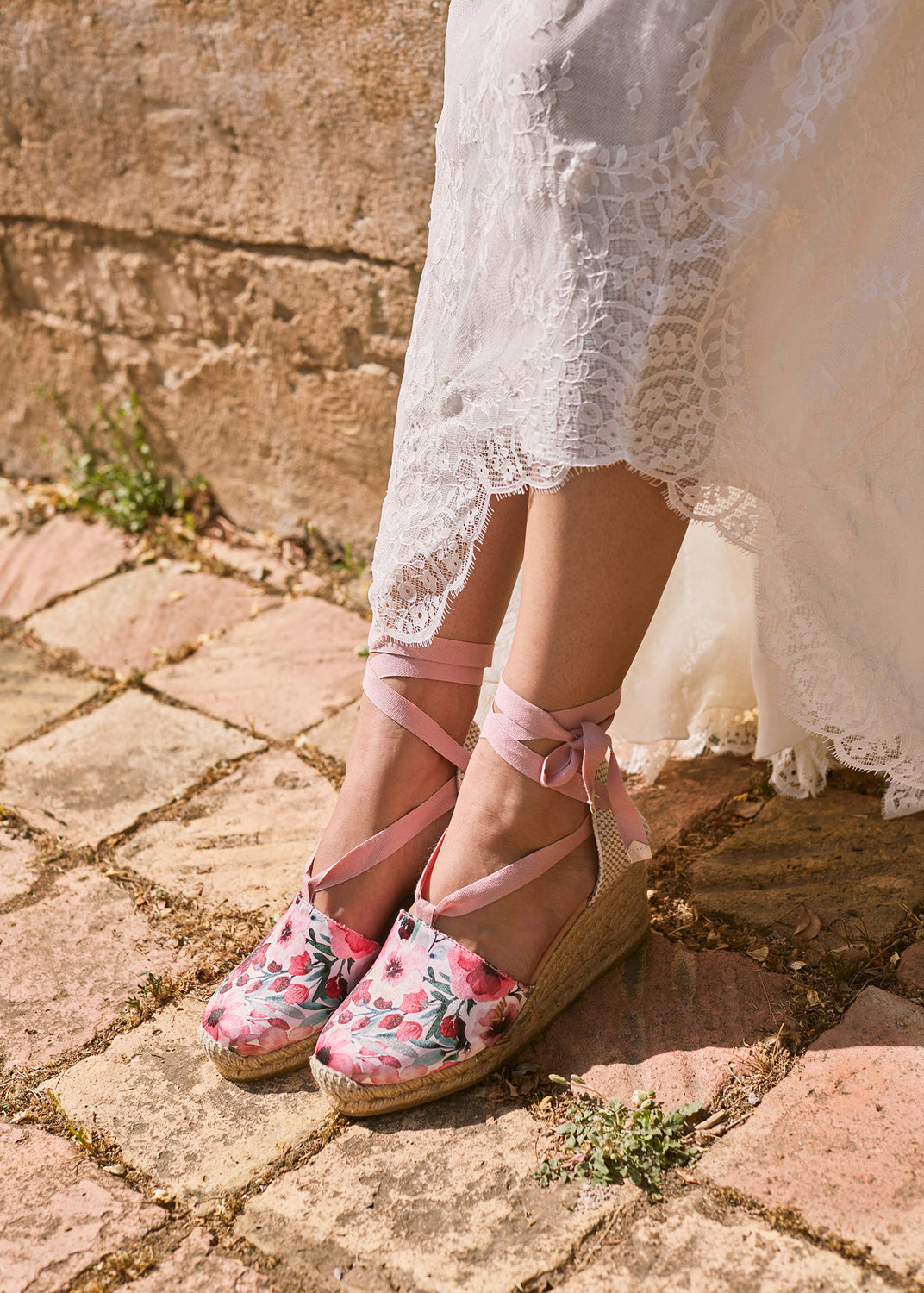 Pink floral espadrille wedge sandals with ribbon ankle ties styled beneath a white lace dress in warm Mediterranean sunlight.