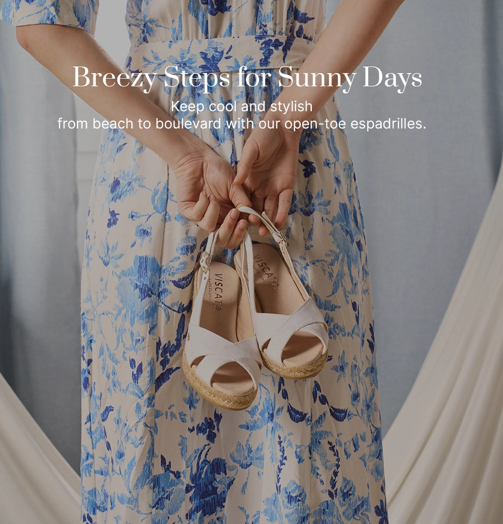 Woman in a blue-and-white floral dress holding a pair of white open-toe espadrille wedges with ankle straps from Viscata, showcasing breathable summer footwear handcrafted in Spain