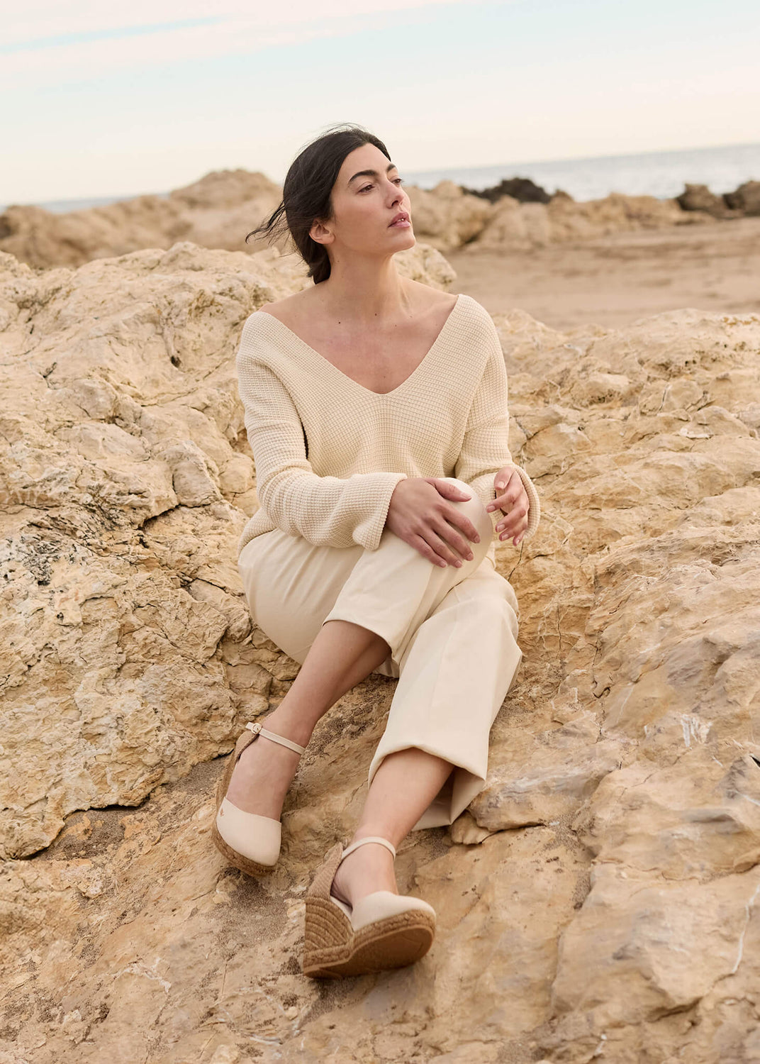 Woman seated on sun-warmed rocks by the coast, dressed in soft neutral tones and wearing Estartit beige canvas ankle-strap wedge espadrilles. The scene captures effortless Mediterranean calm and natural sophistication.