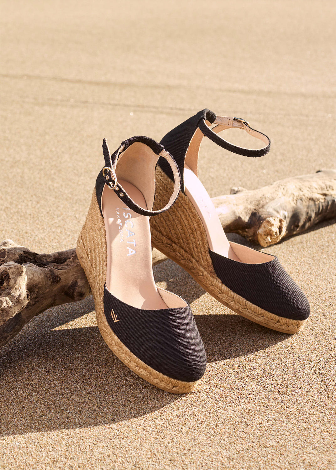 Two black ankle-strap espadrille wedges with closed rounded toes and jute-wrapped wedge soles stand on beach sand beside driftwood.