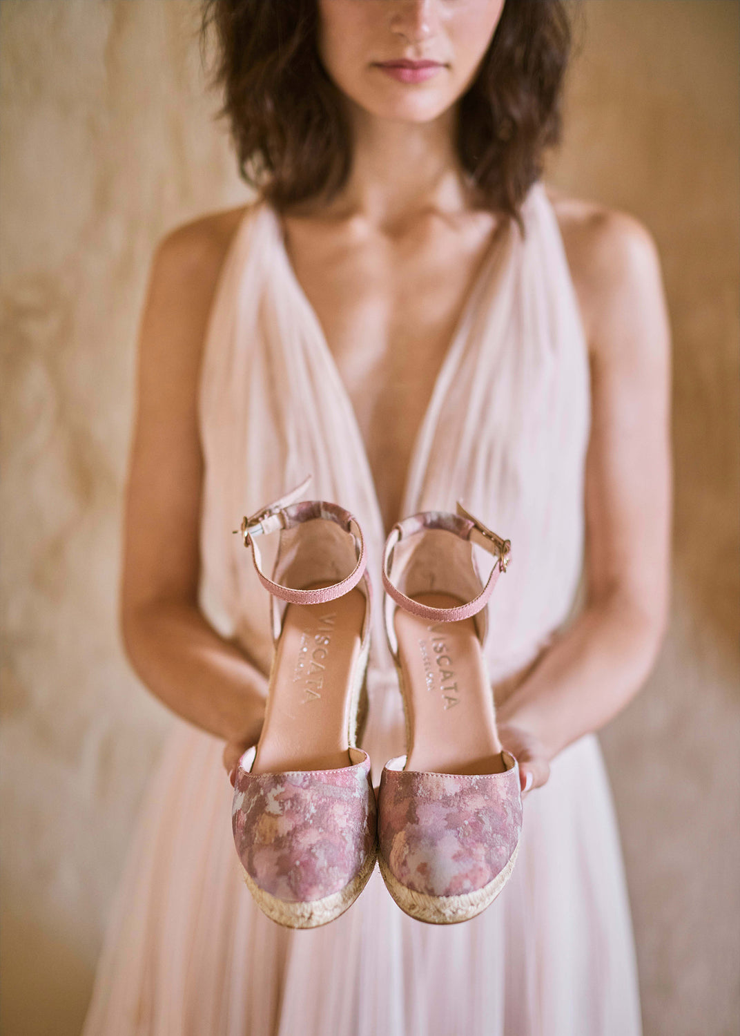 Woman holding pink floral espadrille wedges with ankle straps, styled with a soft blush dress in a warm Mediterranean interior.