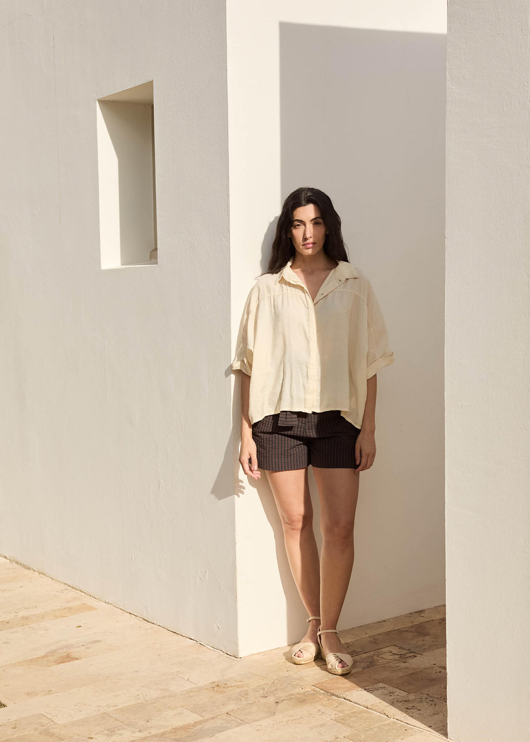 Woman standing against a sunlit white wall wearing a loose beige shirt, dark shorts, and beige ankle-strap espadrille wedges.