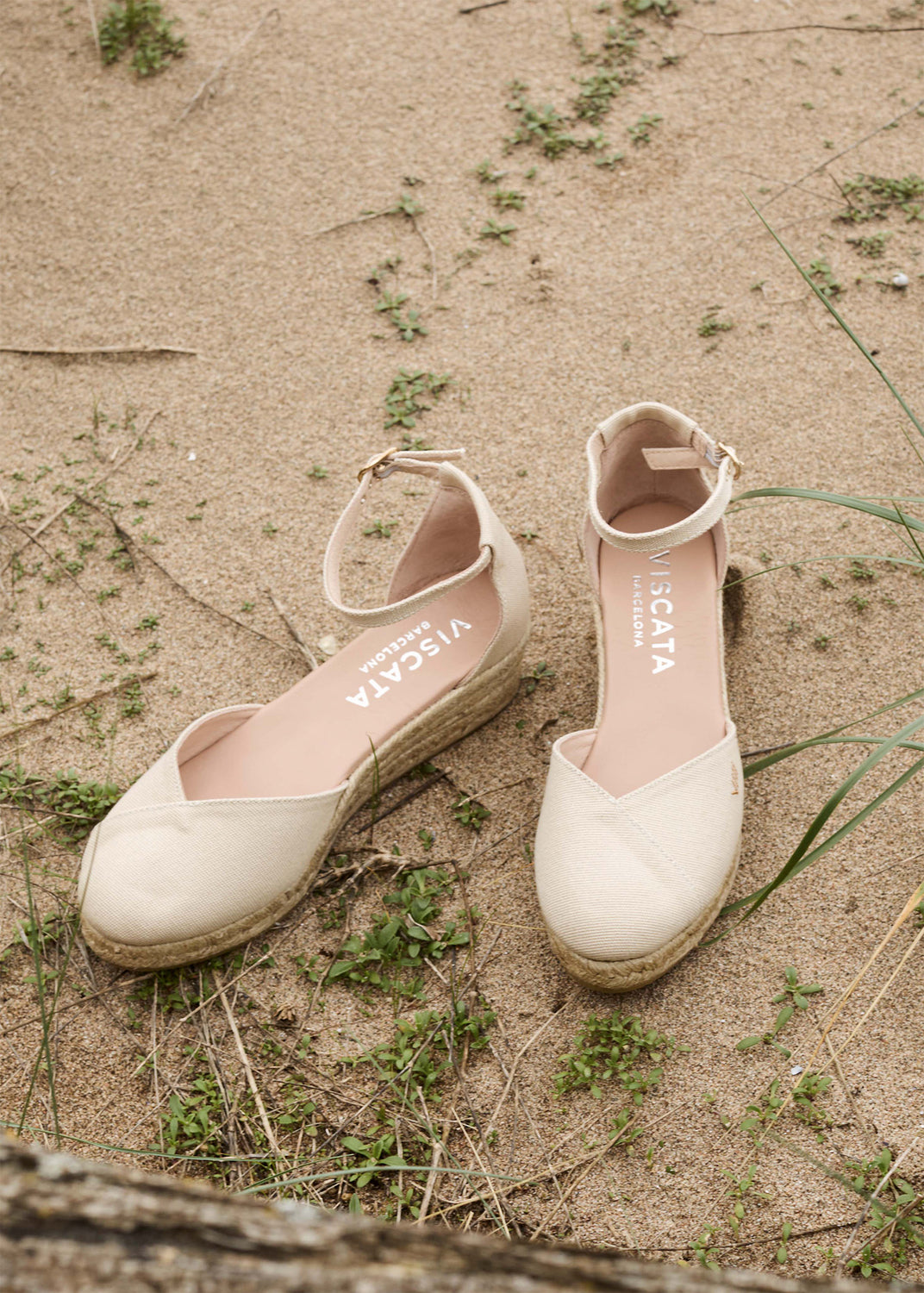 Two beige ankle-strap espadrille flats with closed rounded toes and jute-wrapped soles rest on sandy ground with scattered grass.