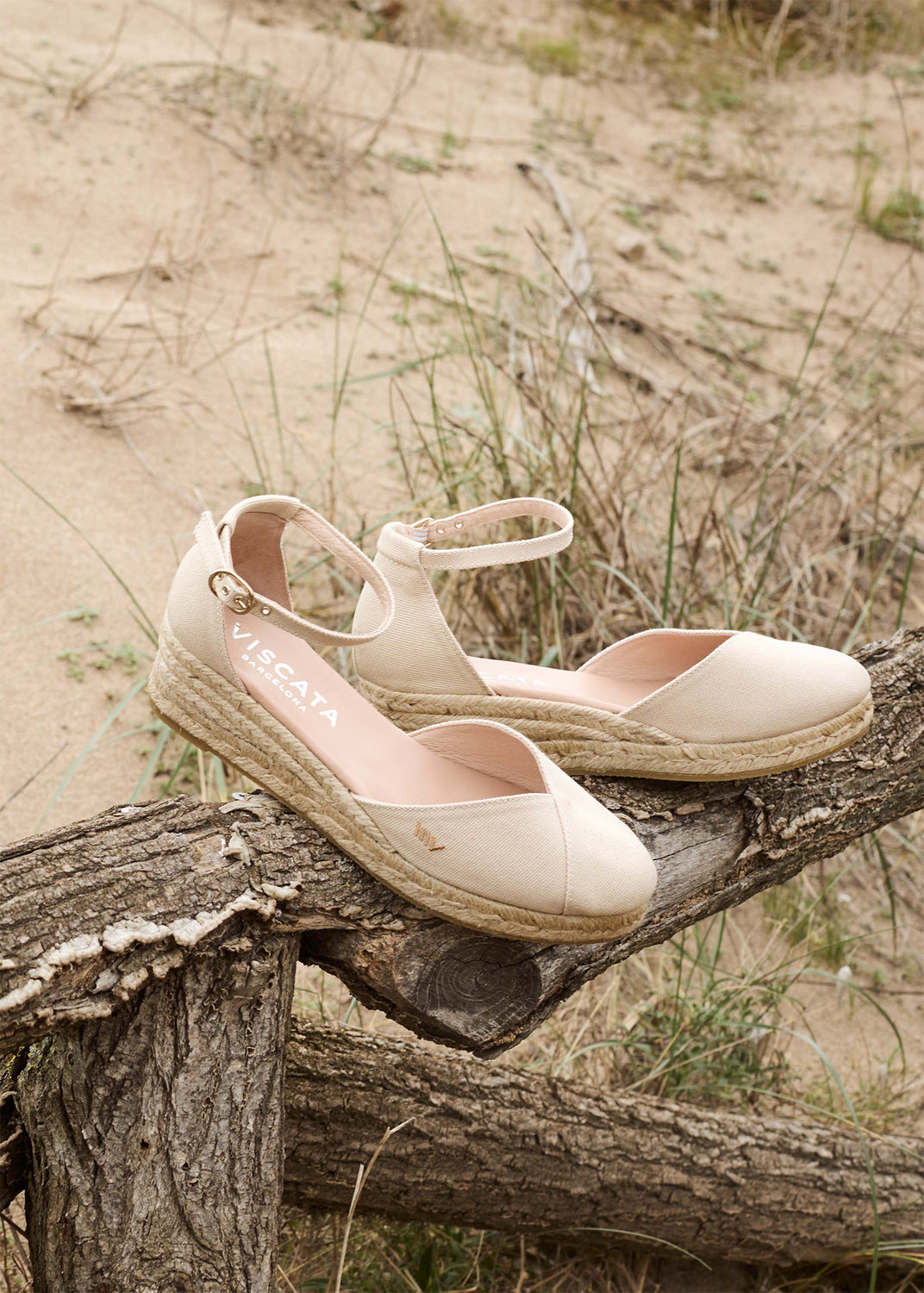 Beige ankle-strap espadrille flats with closed rounded toes and jute-wrapped soles rest on a weathered wooden fence in sandy dunes.