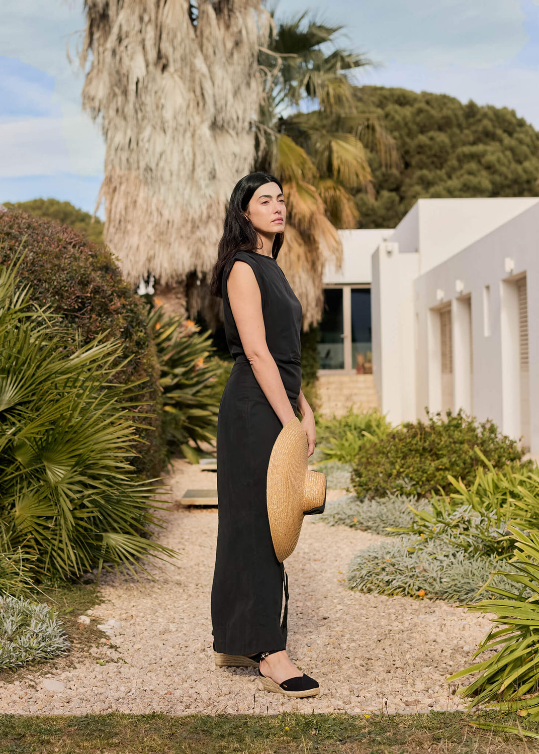 Woman standing along a sunlit garden path lined with palms, wearing a black sleeveless dress and Formiga black ankle-strap wedge espadrilles. She holds a wide straw hat, embodying effortless Mediterranean sophistication.