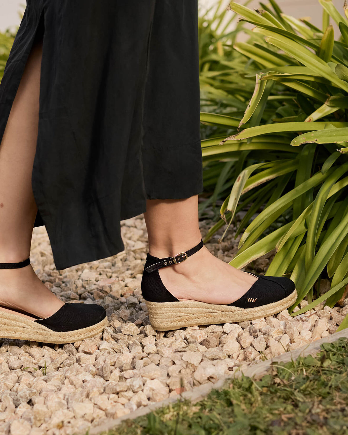 Close-up of a woman walking on a stone path surrounded by greenery, wearing Formiga black canvas ankle-strap wedge espadrilles paired with a flowing black skirt, evoking natural grace and timeless simplicity.